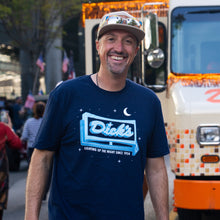 Load image into Gallery viewer, Man wearing the blue crew-neck Lighting Up the Night t-shirt with a logo and text, standing in front of a vehicle.