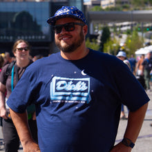 Load image into Gallery viewer, Man wearing the blue crew-neck Lighting Up the Night glow-in-the-dark T-shirt with the Dick's pylon sign, wearing sunglasses and a Dick's Drive-In hat.