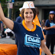Load image into Gallery viewer, Woman wearing the blue V-neck Lighting Up the Night t-shirt with a logo and text, wearing an orange necklace, and a white hat with orange accents.
